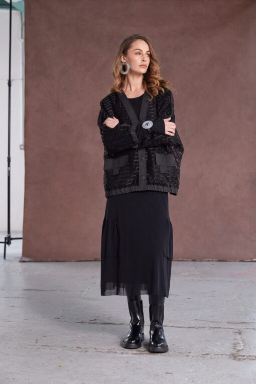 Model wearing NAYA round neck dress with mesh skirt and frill hem (NAW25277), layered with a black textured jacket and paired with patent ankle boots, standing against a brown backdrop.
