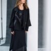 Model wearing NAYA black angled hem skirt (NAW25278) paired with a layered black top and relaxed black jacket, styled with chunky boots, standing in a minimal studio space.