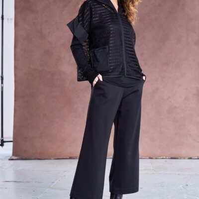 Model wearing black NAYA cropped wide leg trousers with a textured black zip-up jacket and patent ankle boots, standing against a soft brown backdrop.