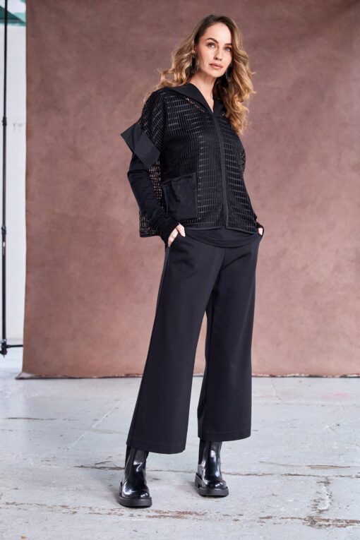 Model wearing black NAYA cropped wide leg trousers with a textured black zip-up jacket and patent ankle boots, standing against a soft brown backdrop.