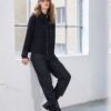 Model wearing NAYA pinstriped pull-up trousers with side pockets and a black zip-up jacket, leaning casually against a white wall in a bright studio setting.