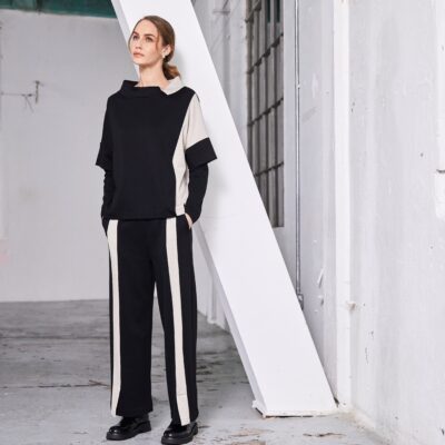 Model wearing NAYA wide leg black trousers with sand contrast panel (NAW25264), paired with a matching black top with side stripe, standing in a minimalist industrial setting.