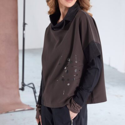 Model wearing NAYA placement print top with contrast cut-away collar (NAW25276), styled with black cargo trousers and a black cap, smiling in a studio setting.