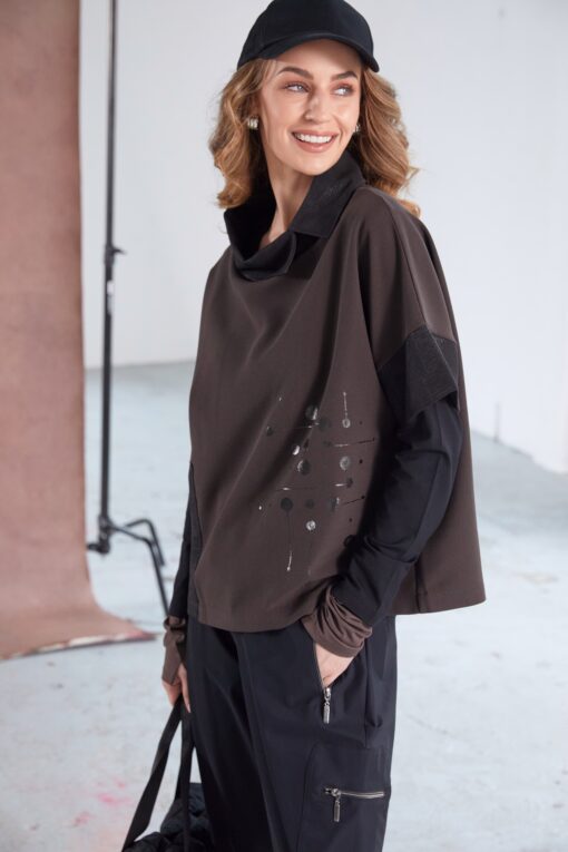 Model wearing NAYA placement print top with contrast cut-away collar (NAW25276), styled with black cargo trousers and a black cap, smiling in a studio setting.