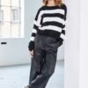 Model wearing NAYA wide leg faux leather trousers paired with a black and white striped jumper and black sneakers, leaning casually against a white wall in a bright studio.