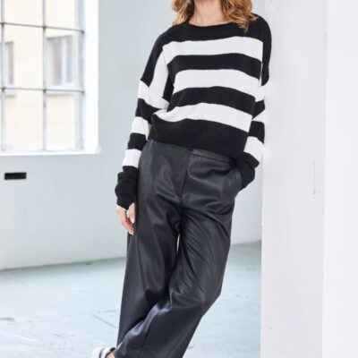 Model wearing NAYA wide leg faux leather trousers paired with a black and white striped jumper and black sneakers, leaning casually against a white wall in a bright studio.