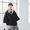 Model wearing black and taupe NAYA high neck jacket with wide sleeves and stud closure, styled with patterned trousers and a black cap, standing by a window.