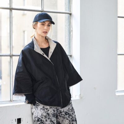 Model wearing black and taupe NAYA high neck jacket with wide sleeves and stud closure, styled with patterned trousers and a black cap, standing by a window.